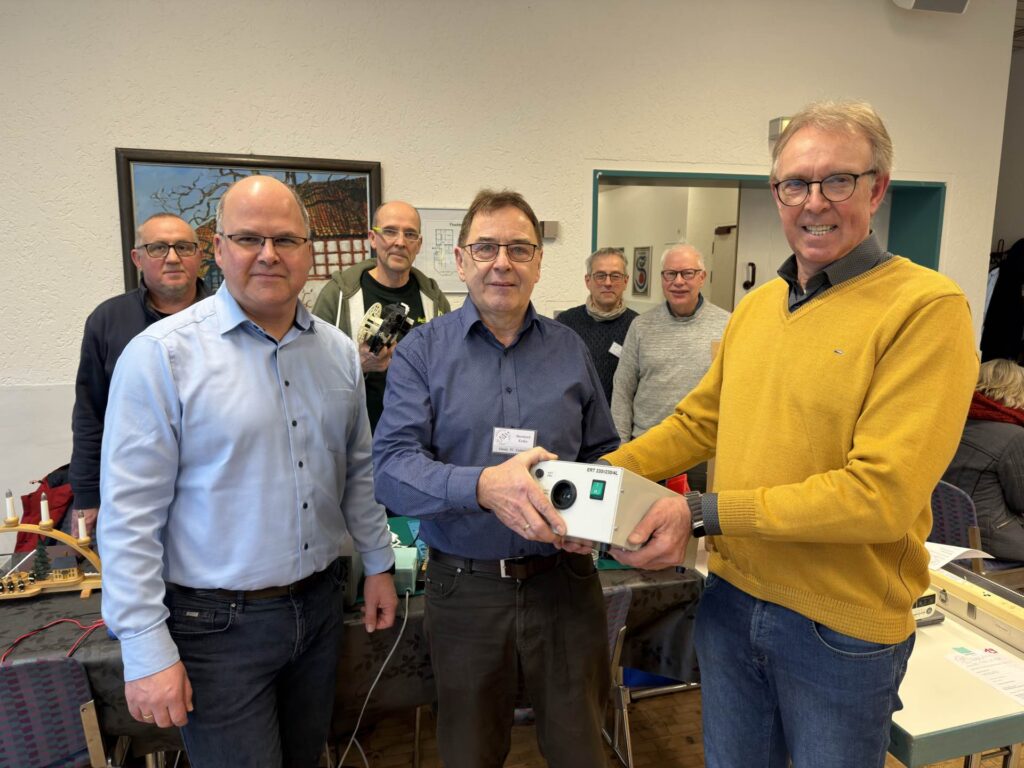 Dr. Eberhard Niggemann (Vorsitzender der Bürgerstiftung Schlangen) übergibt im Beisein von Bürgermeister Marcus Püster den neuen Trenntransformator an Bernhard Krake vom Reparatur-Café. Im Hintergrund freuen sich weitere ehrenamtliche Helferinnen und Helfer vom Reparatur-Café über die Unterstützung